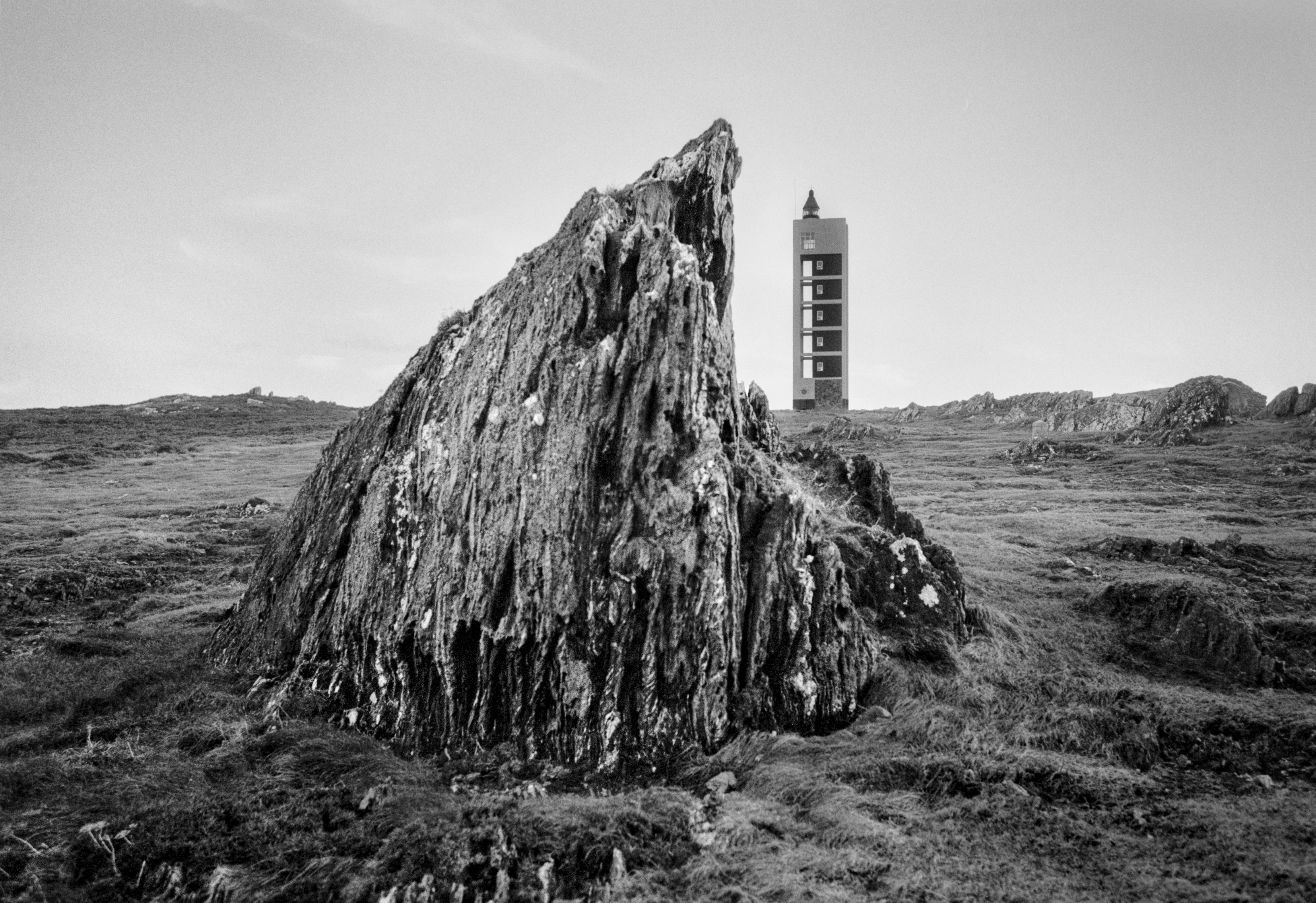 A black and white photograph of a large weathered rock in the foreground with a tall lighthouse standing in the distance on an open coastal landscape under a clear sky.