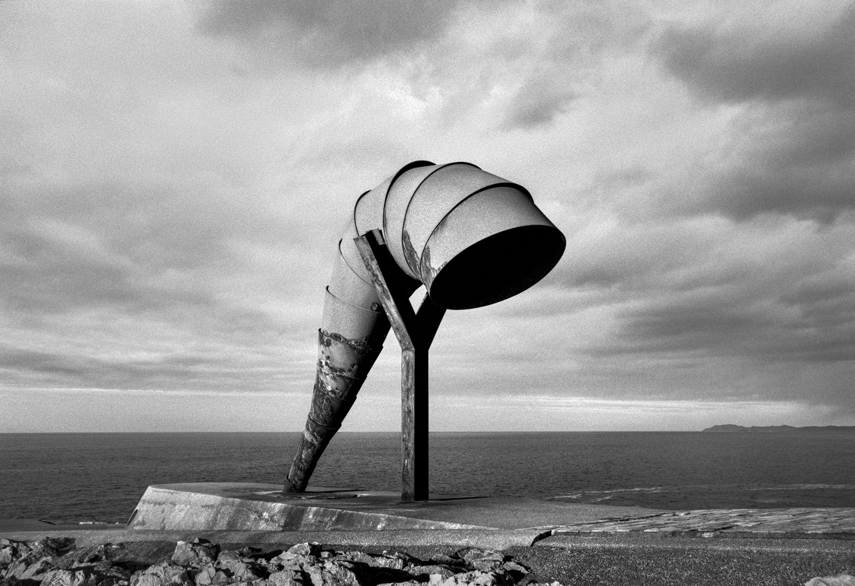 A black and white photograph of a large metal sculpture shaped like a bent horn standing on a coastal platform, with the ocean and a cloudy sky in the background.