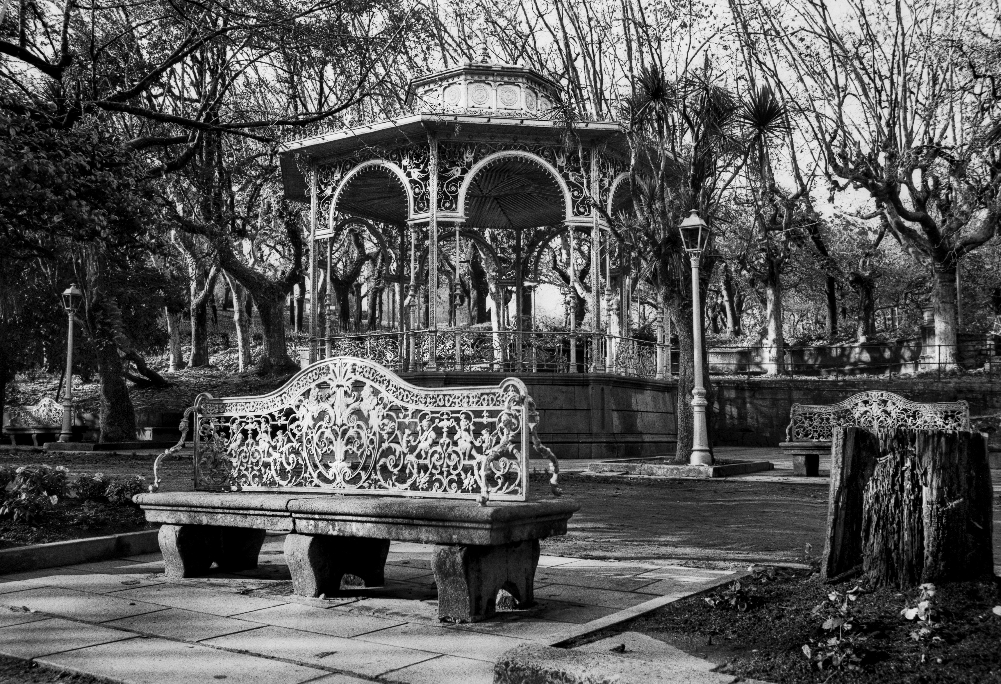 A black and white photograph of an ornate park bench and an elaborate bandstand surrounded by leafless trees in a quiet public garden.