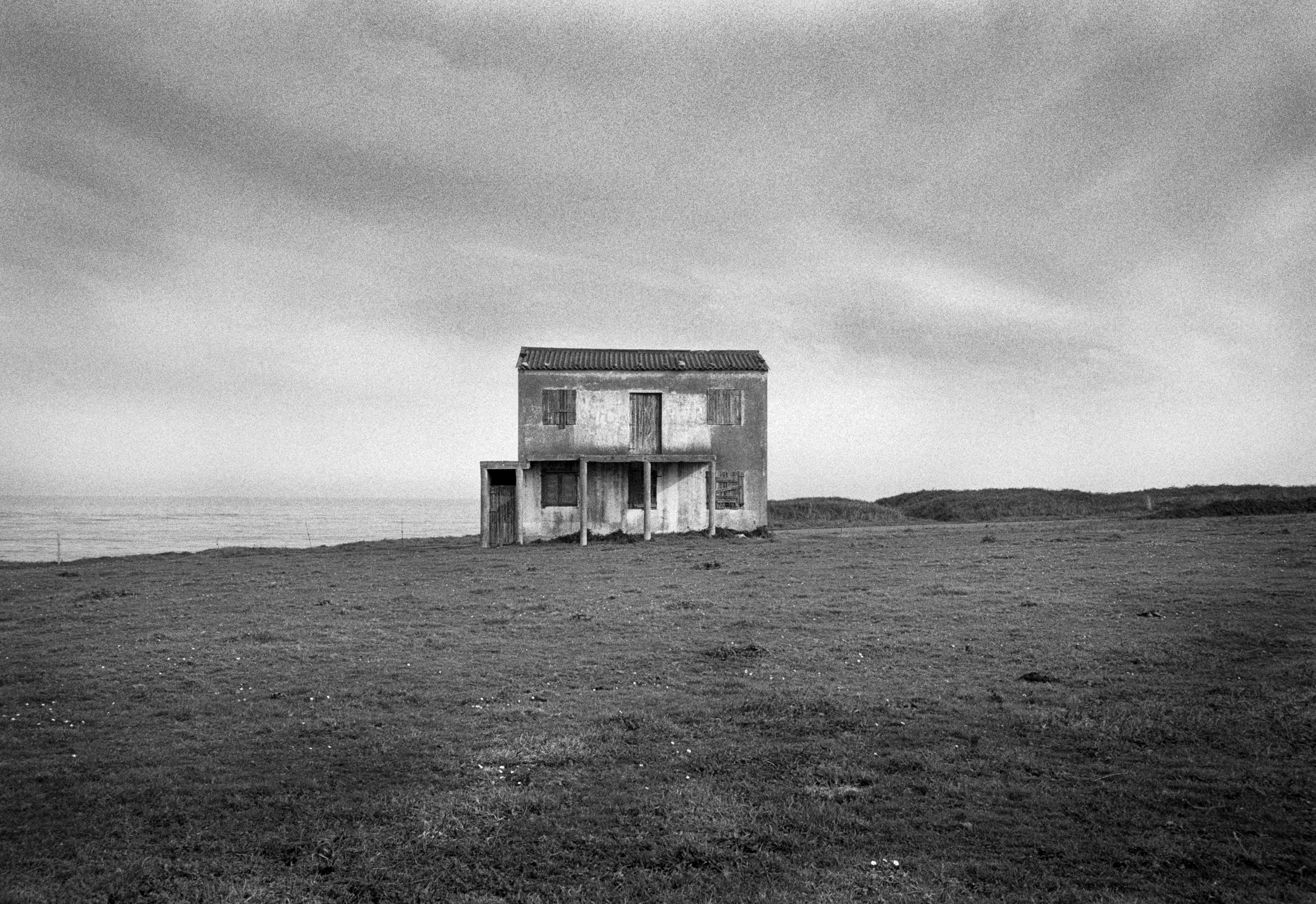 A black and white photograph of an abandoned two-story house standing alone in an open field near the coast, with the sea and cloudy sky in the background.