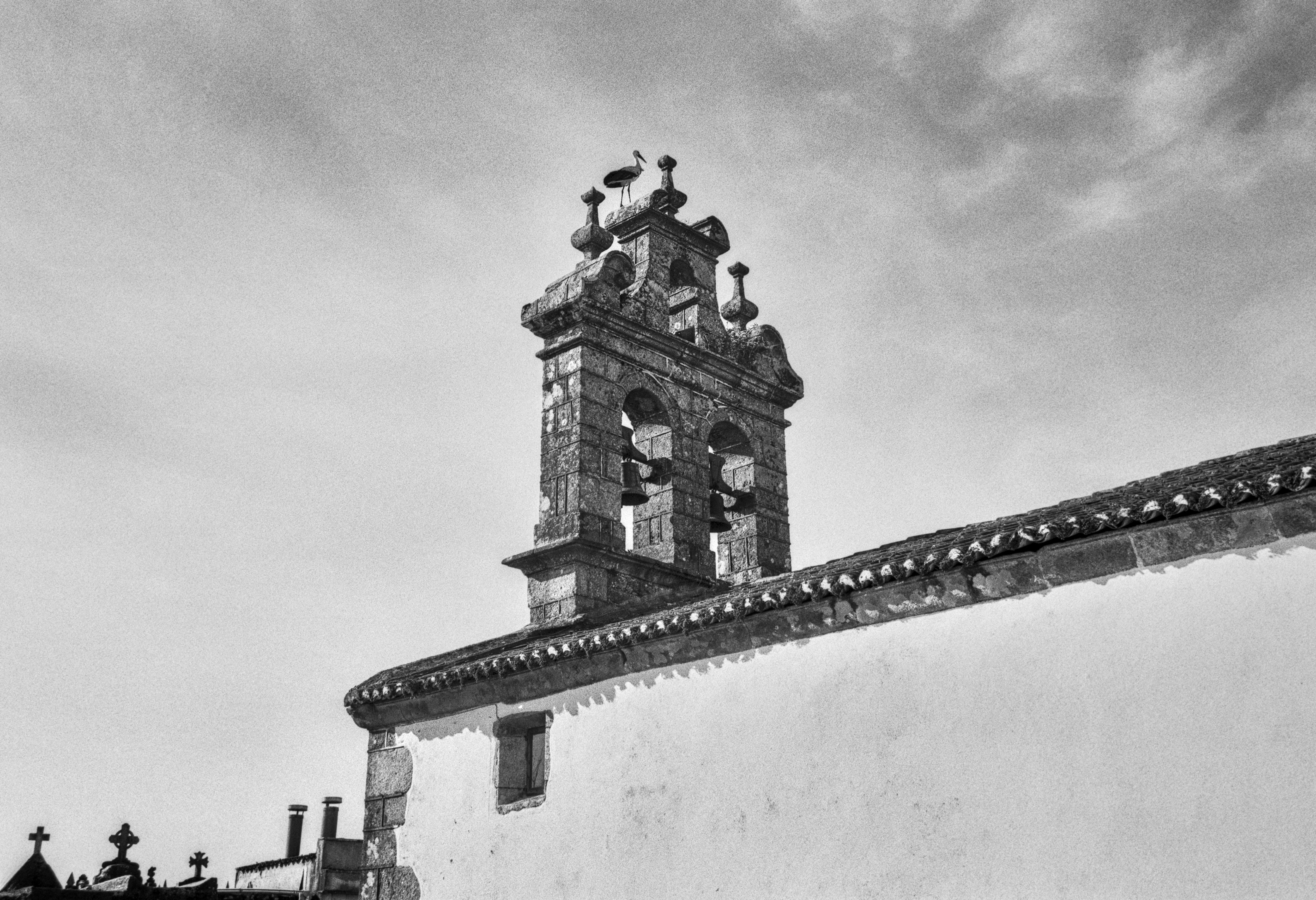 A black and white photograph of a stone church bell tower with two bells and a stork standing on top, seen against a cloudy sky. The roof and part of another building appear in the lower part of the frame.