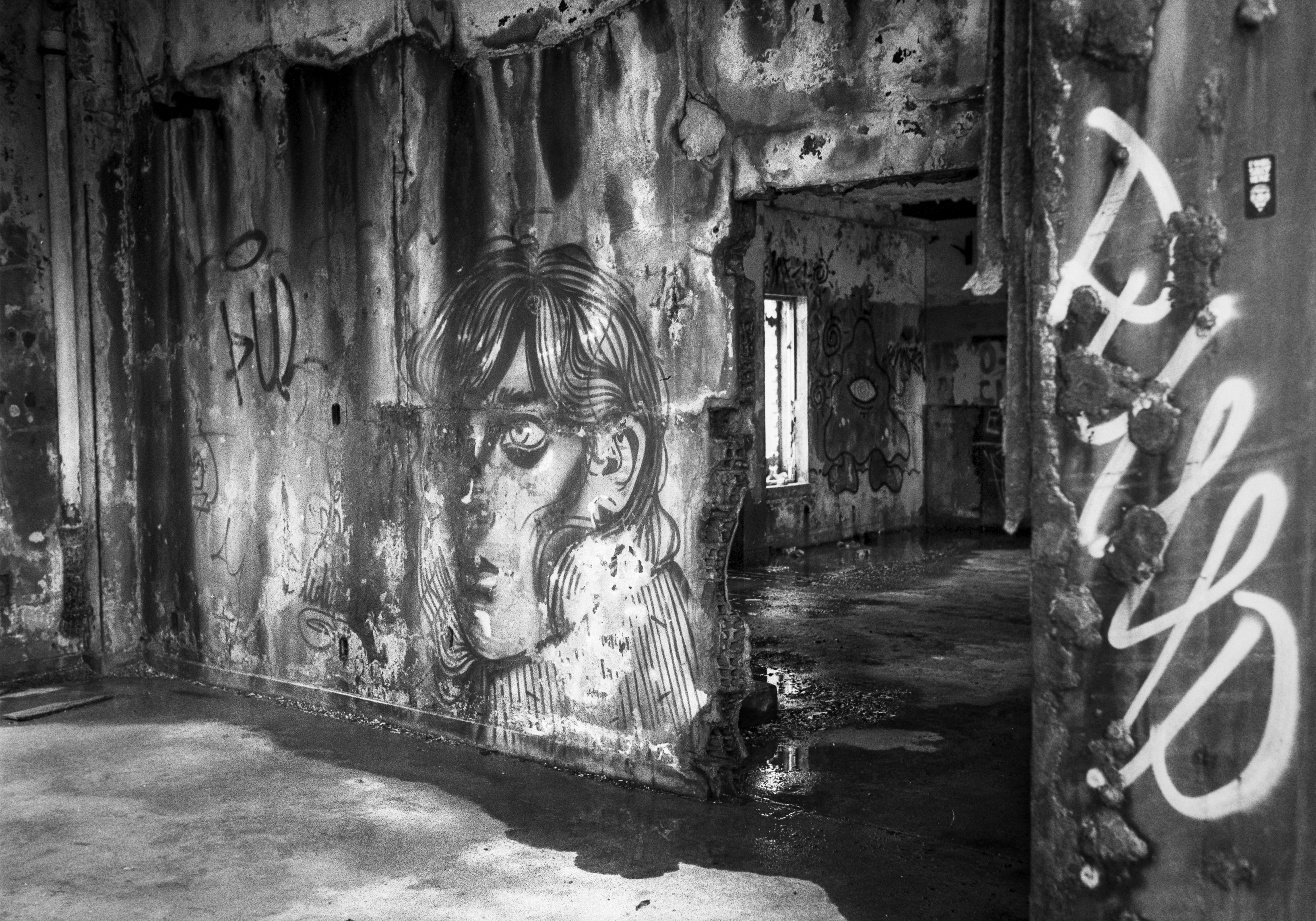 A black and white photograph of an abandoned building interior with damaged walls covered in graffiti, including a large painted face near a doorway.