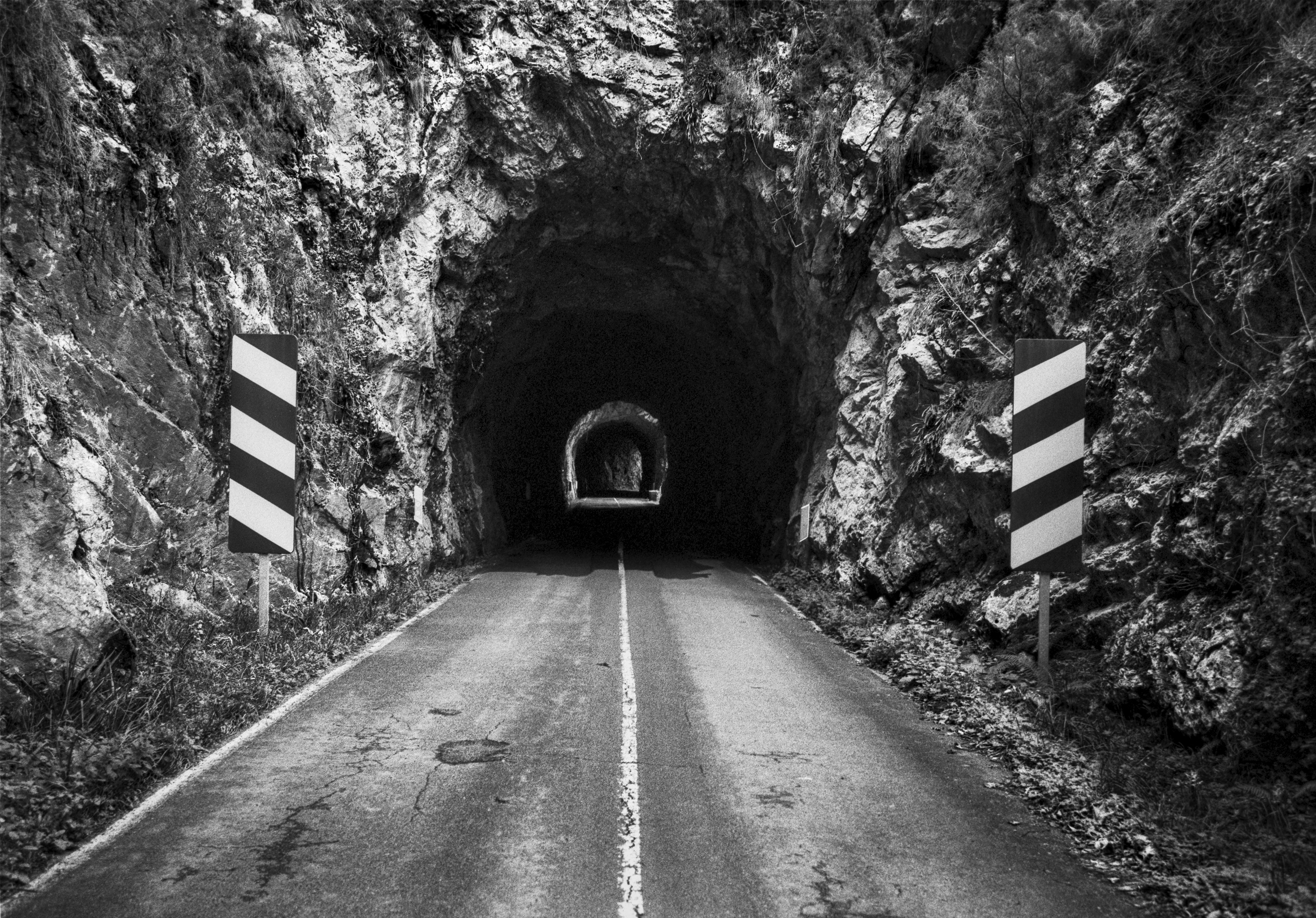 A black and white photograph of a narrow mountain road entering a rock carved tunnel, with a second tunnel visible beyond it and striped warning signs on both sides.