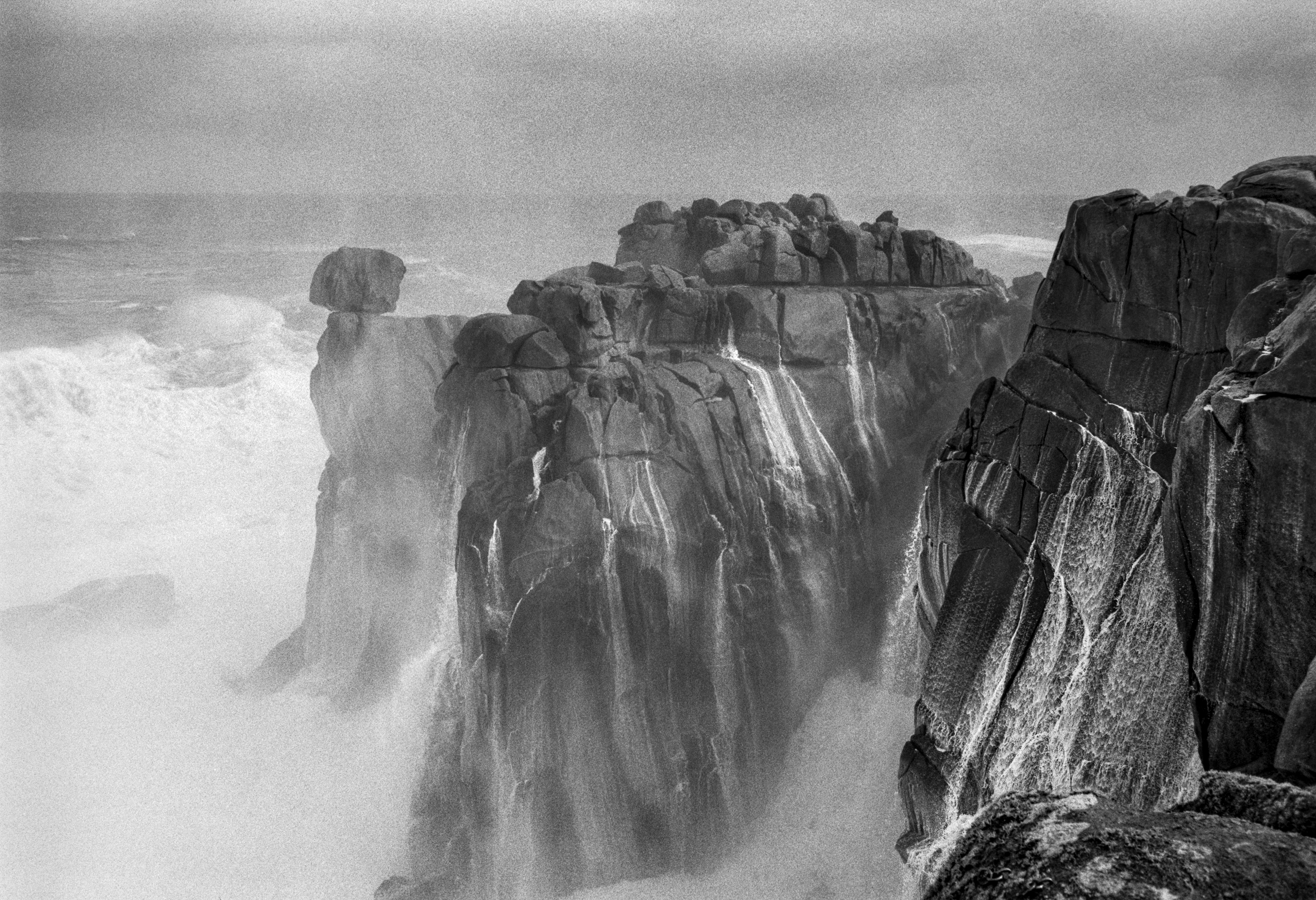 A black and white photograph of massive coastal cliffs with rough rock formations and waves crashing below, partially obscured by sea mist.