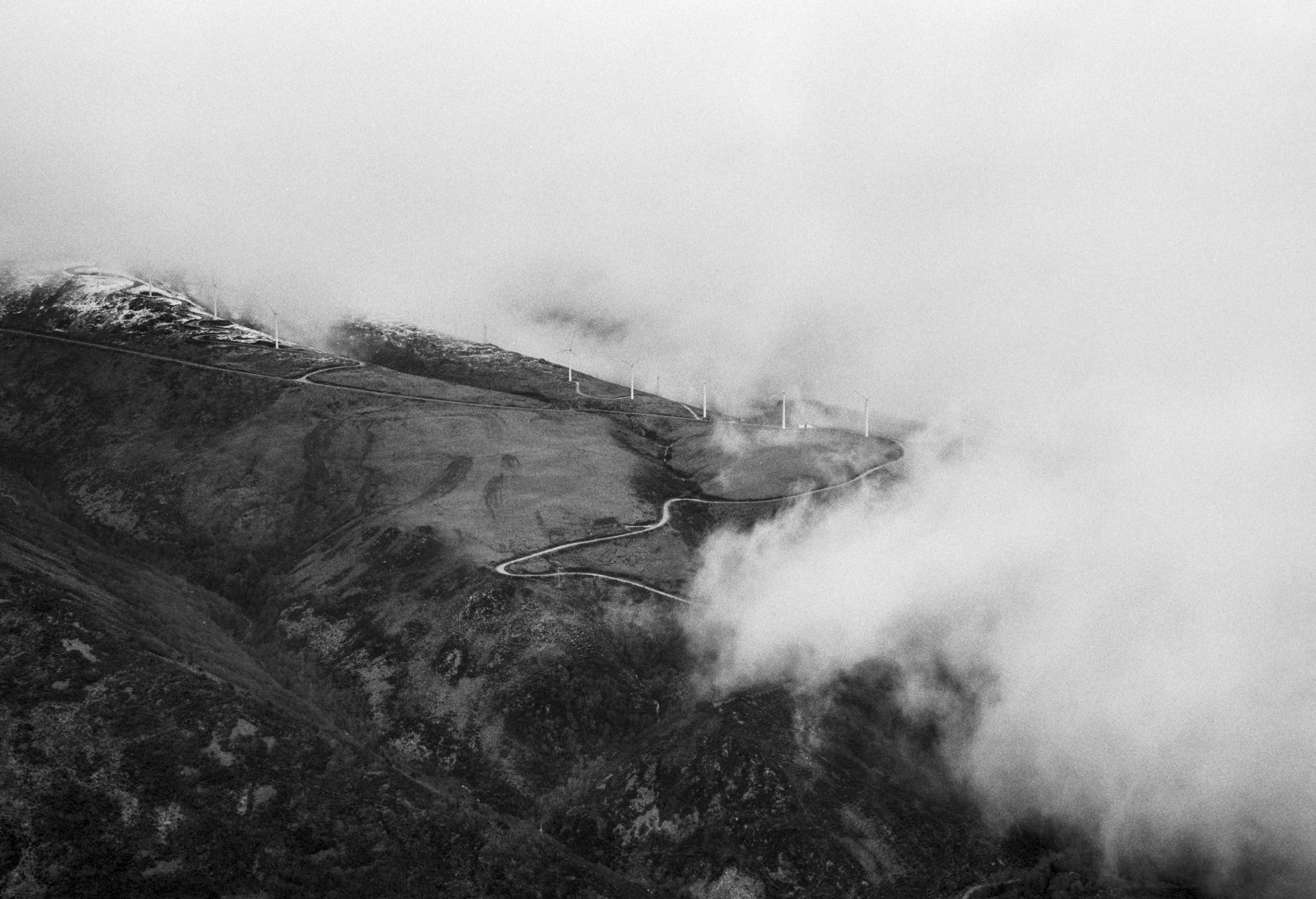 A black and white photograph of a mountain ridge partly covered by clouds, with a winding road and a line of wind turbines visible along the slope.