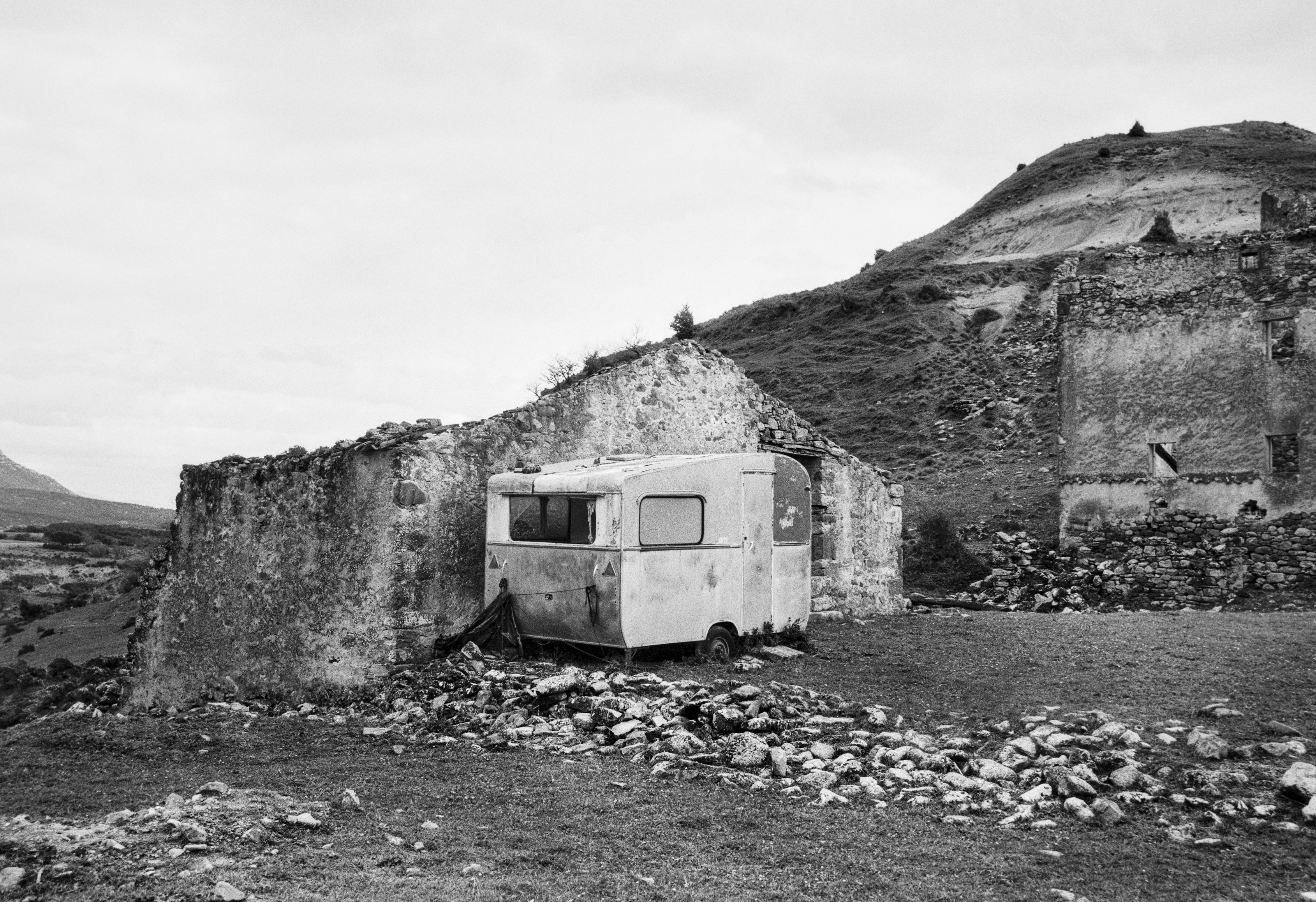 A black and white photograph of an old caravan resting against the ruined stone walls of an abandoned village, with a hillside rising behind it. The ground is covered with scattered rocks.