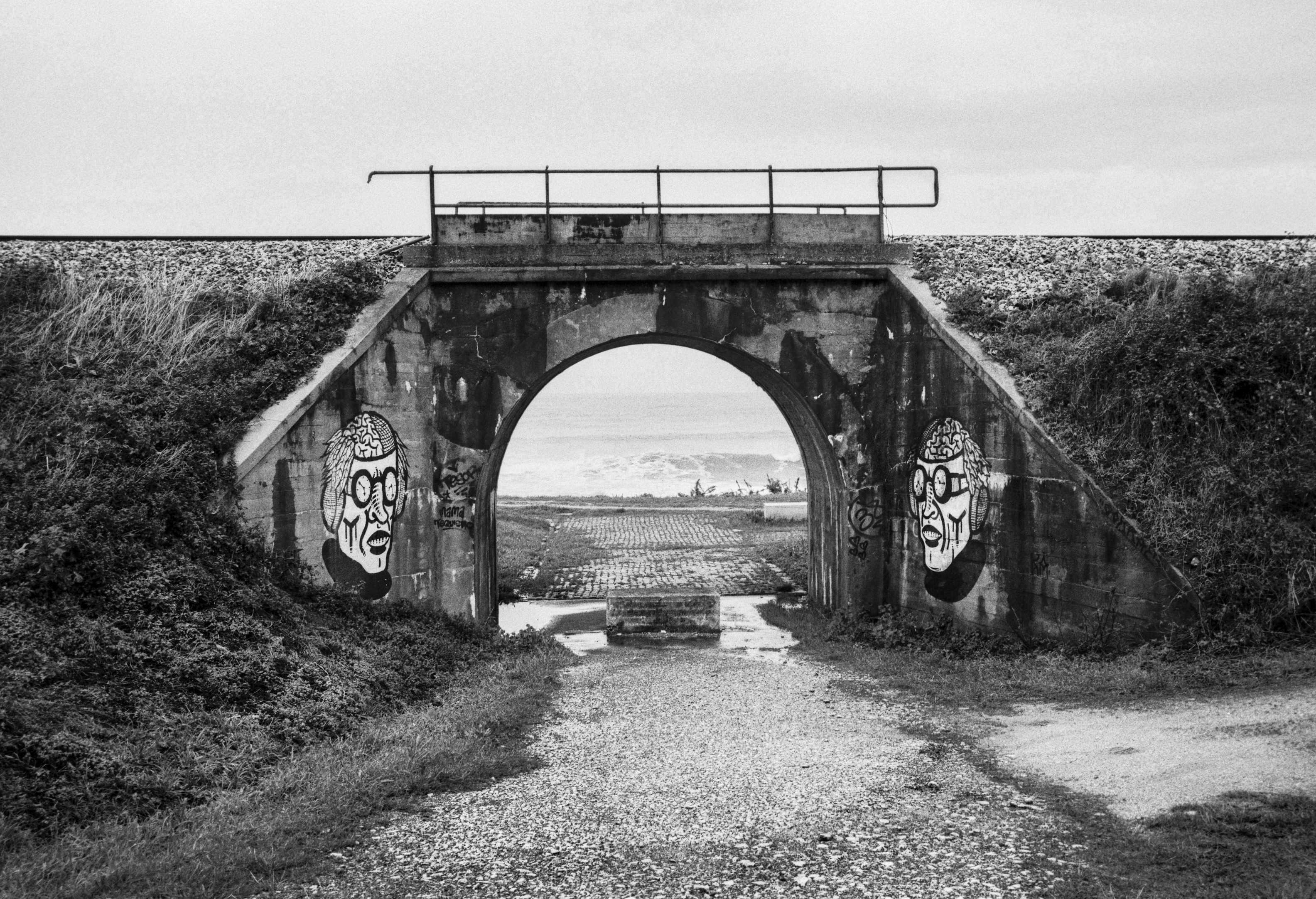 A small railway underpass in A Mariña Lucense, with symmetric graffiti framing a distant view of the sea.