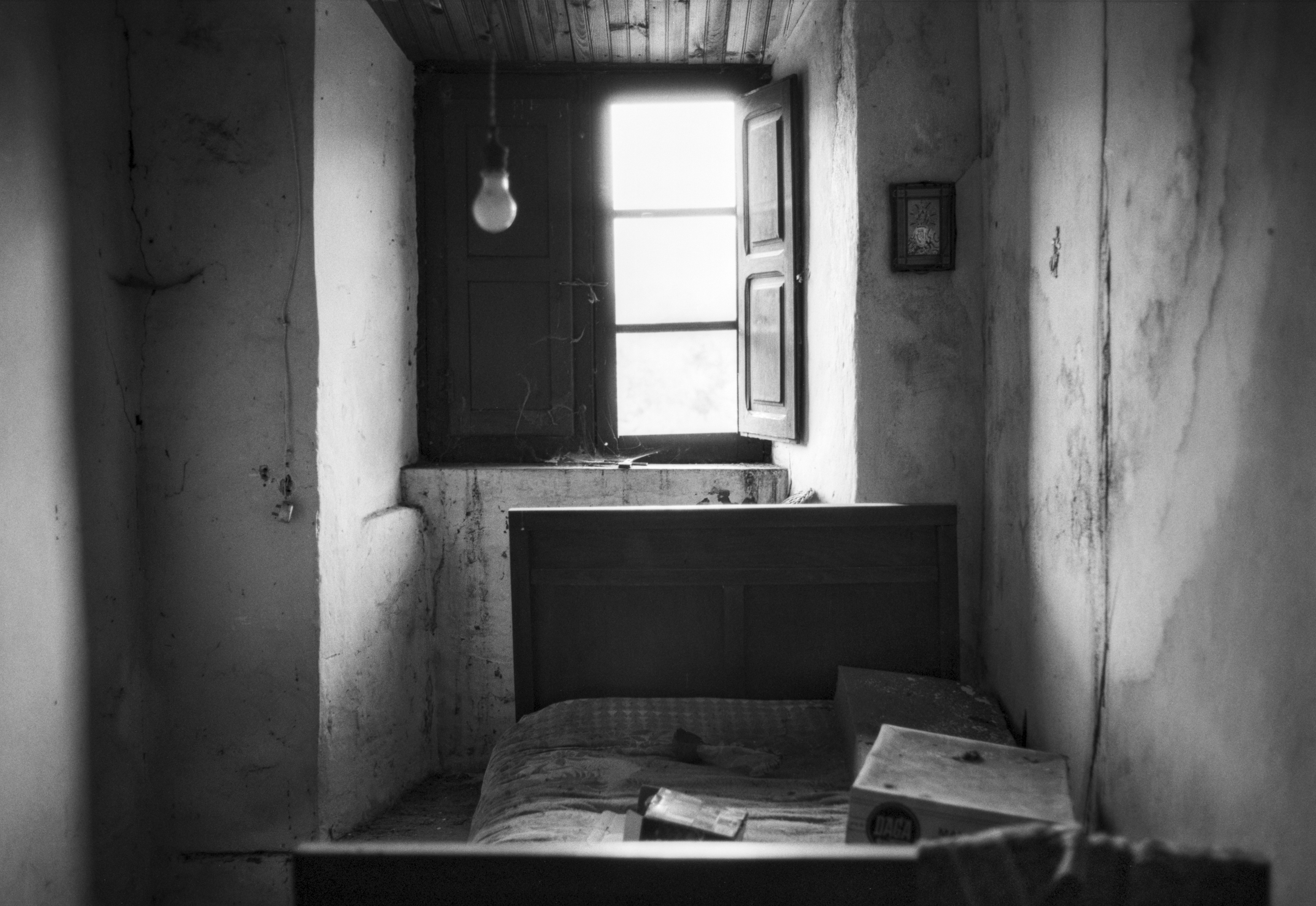 An abandoned rural bedroom with a single bed, a dusty window, and soft light filtering through the open shutters.