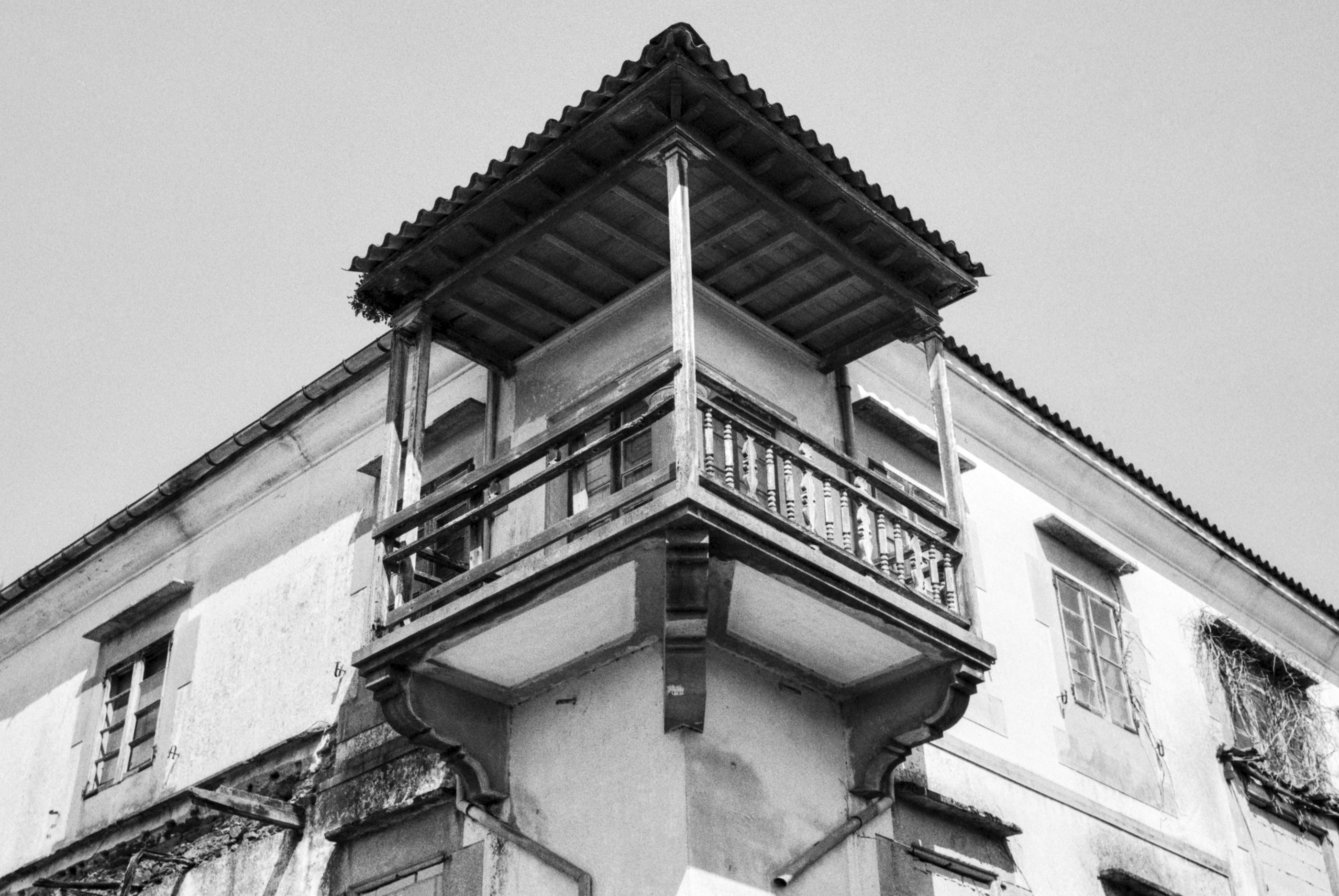 A corner view of an old house with a wooden balcony and tiled roof, photographed from below against an open sky in black and white.